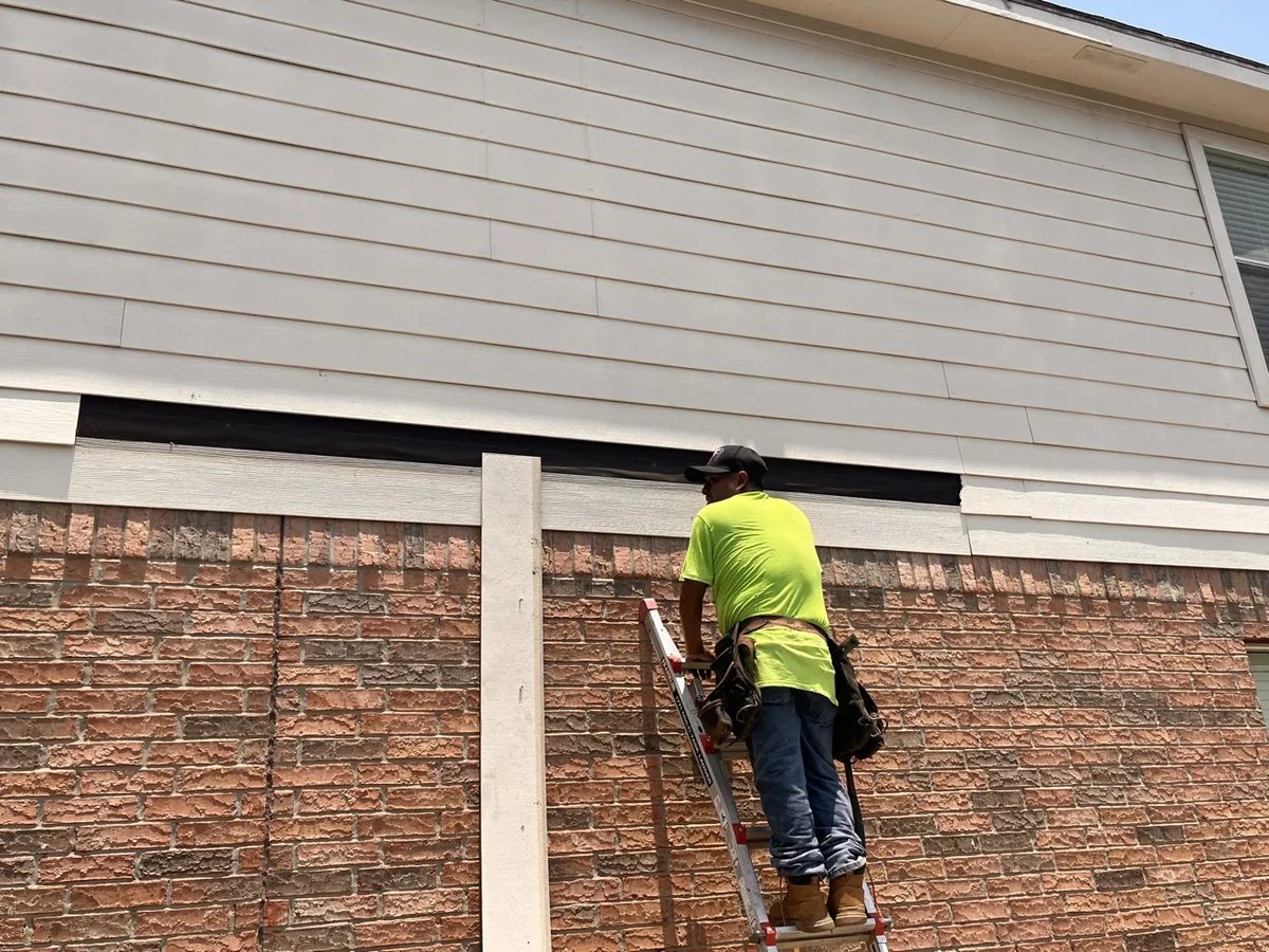 Worker removing damaged fiber-cement siding boards, exposing the weather barrier
